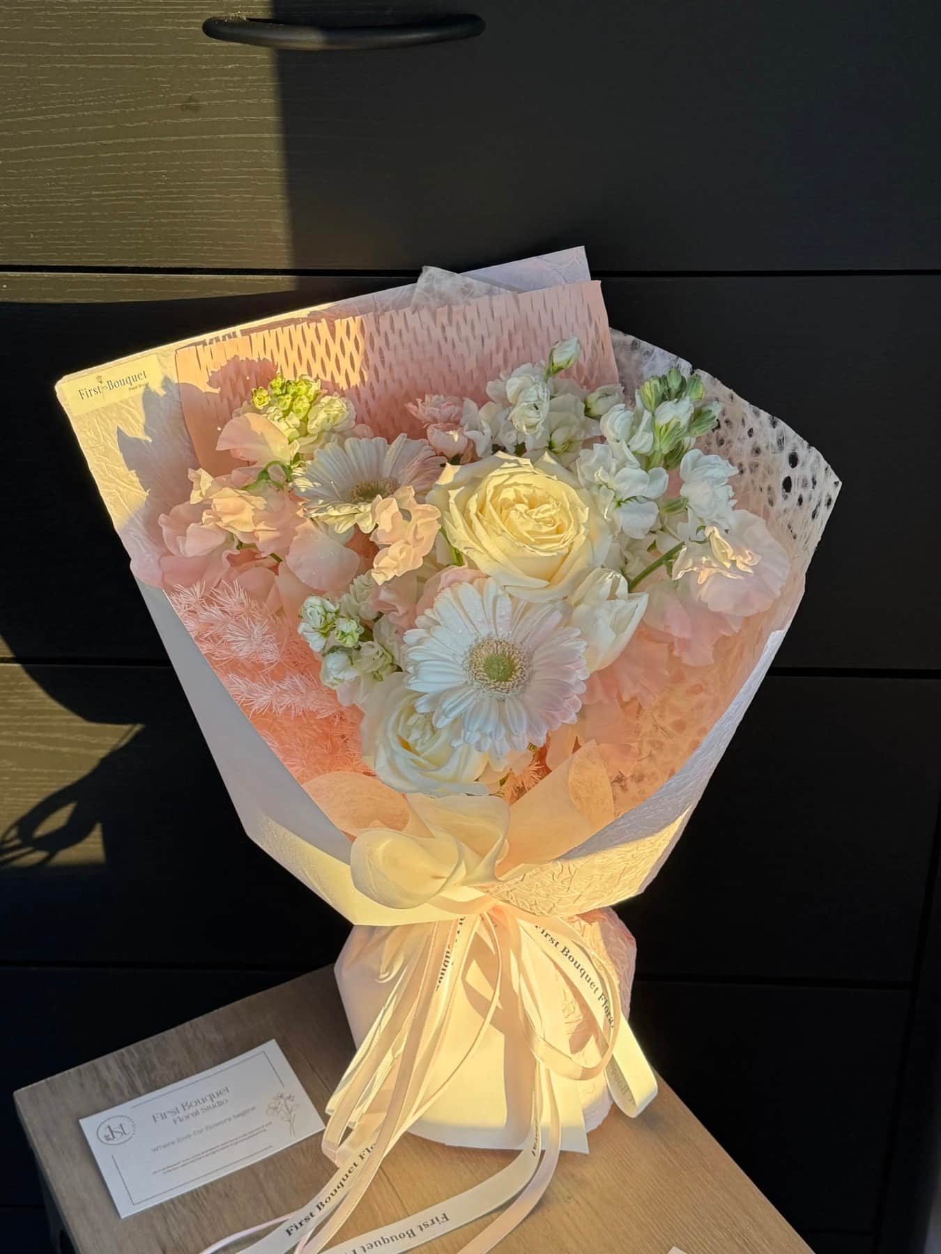 A hand-tied bouquet of white roses, gerbera and stock in blush wrapping, tied with a First Bouquet ribbon, in late sun.