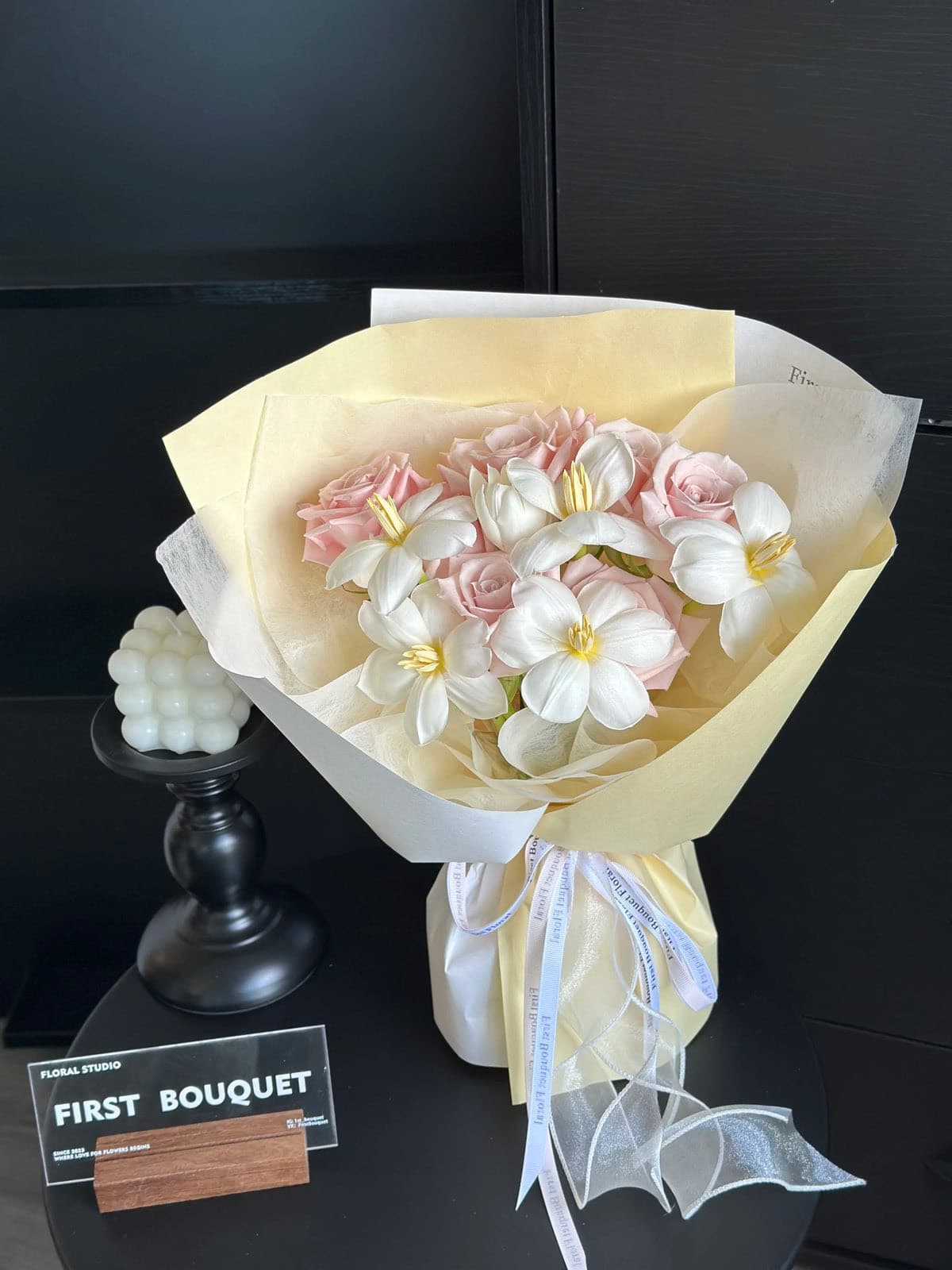 Pink roses and white tulips in pale yellow wrapping, beside a First Bouquet sign.